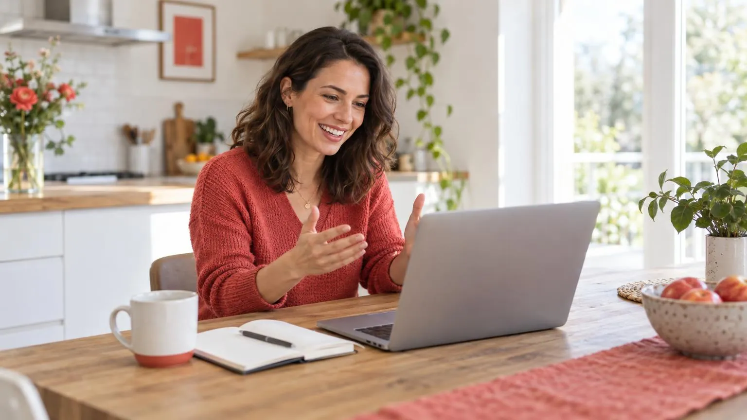A smiling woman talking to someone on a laptop video call at a bright kitchen table